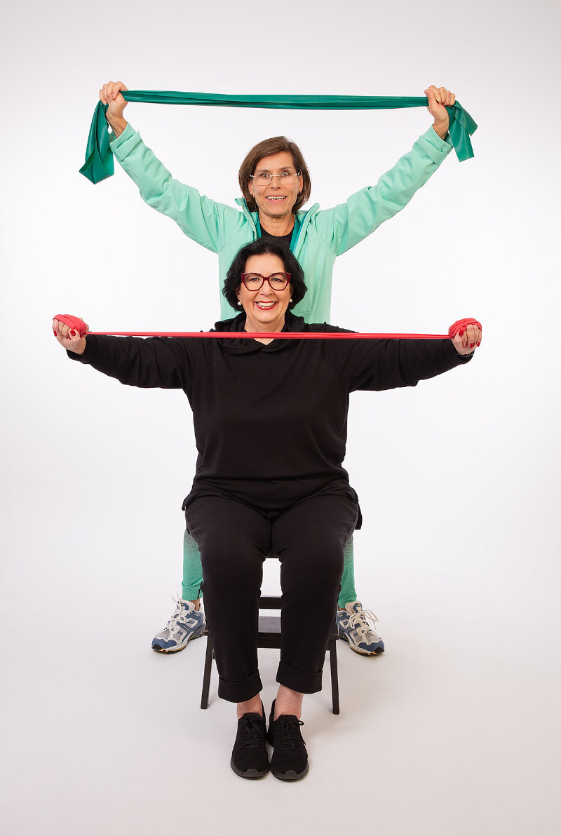 Margitta steht hinter der sitzenden Dagmar und beide halten ein Band ausgestreckt vor sich